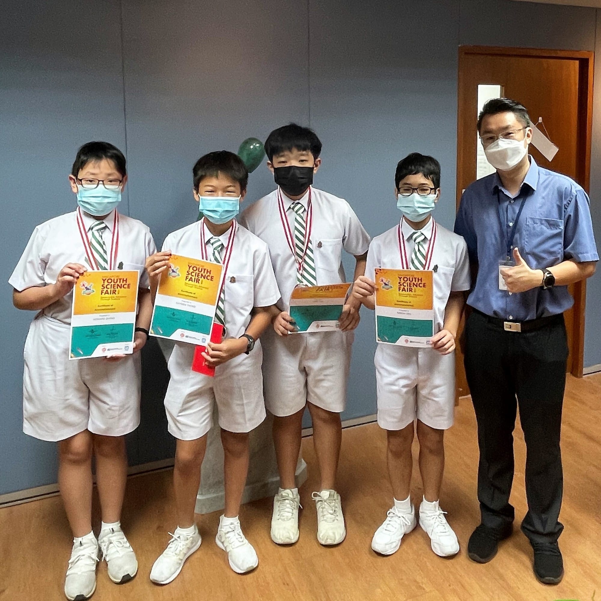 Four masked students in school uniforms hold Youth Science Fair certificates with medals, posing with a masked adult teacher.